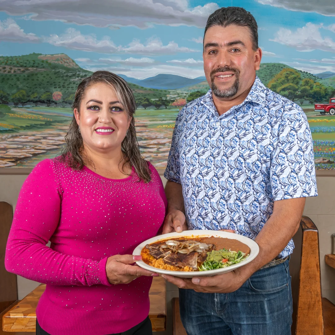 Gerardo Aguirre and Ana Gloria Fernandez smiling together, holding a plate of Mexican food in their restaurant